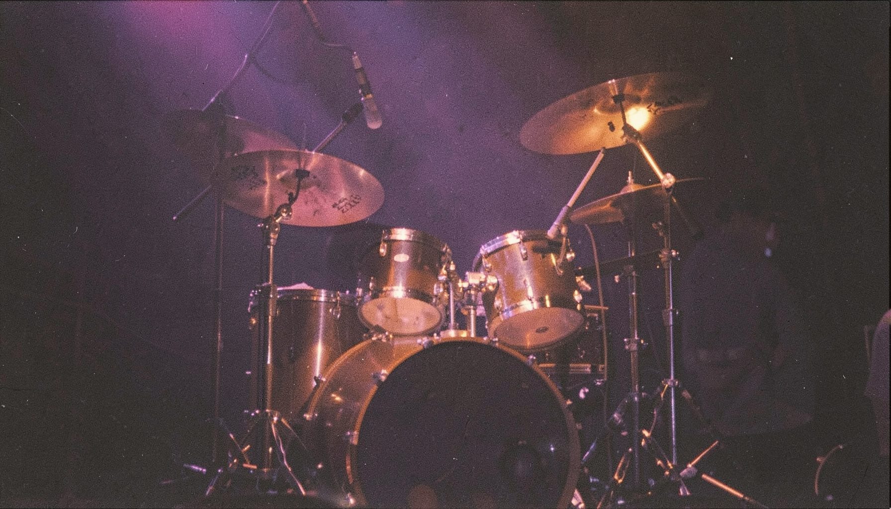 A warm, softly lit analog film photograph with visible grain showing a drum kit on a dark stage, deep violet and amber stage lights casting a dramatic glow across the drums and cymbals.