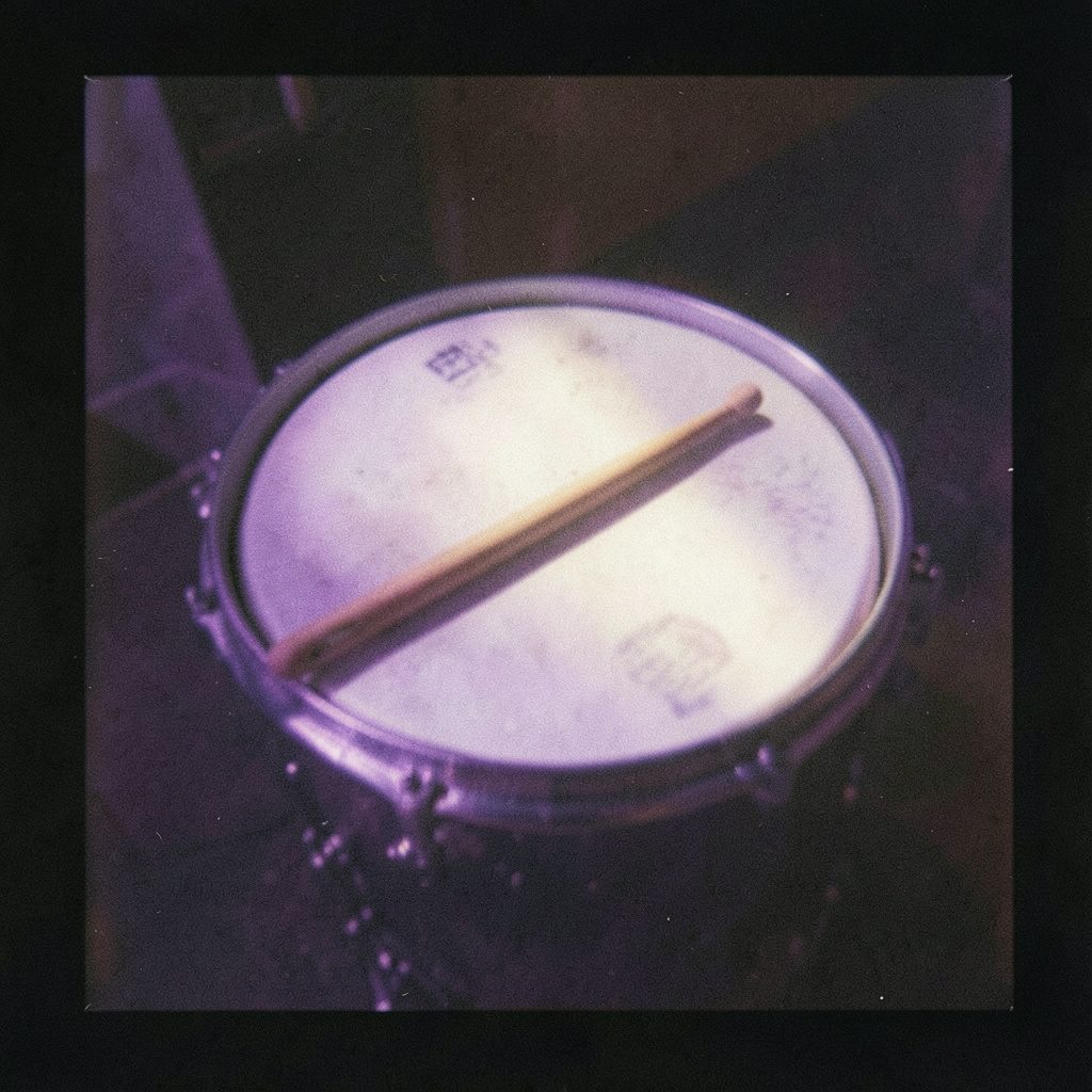 A small square analog film photograph with visible grain, a single drumstick resting diagonally across a drumhead, deep indigo shadows and warm amber rim light creating a quiet, contemplative mood.