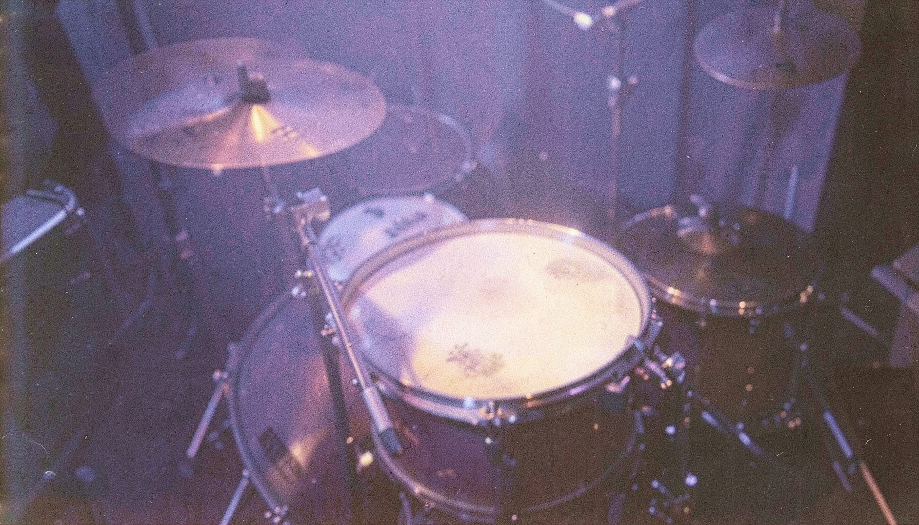 A grainy analog film photograph of a snare drum and brushed cymbals in soft lavender-blue stage light, warm cream highlights catching the metal rims and drumhead texture.